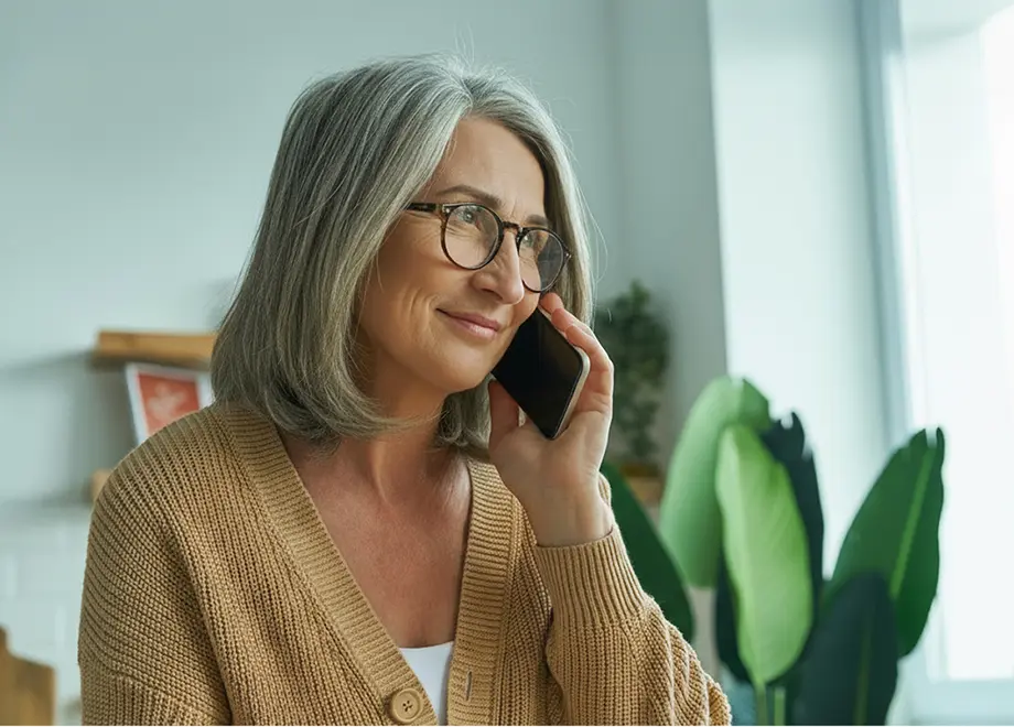 Smiling middle-aged woman with gray hair and glasses talking on a smartphone indoors near green plants.
