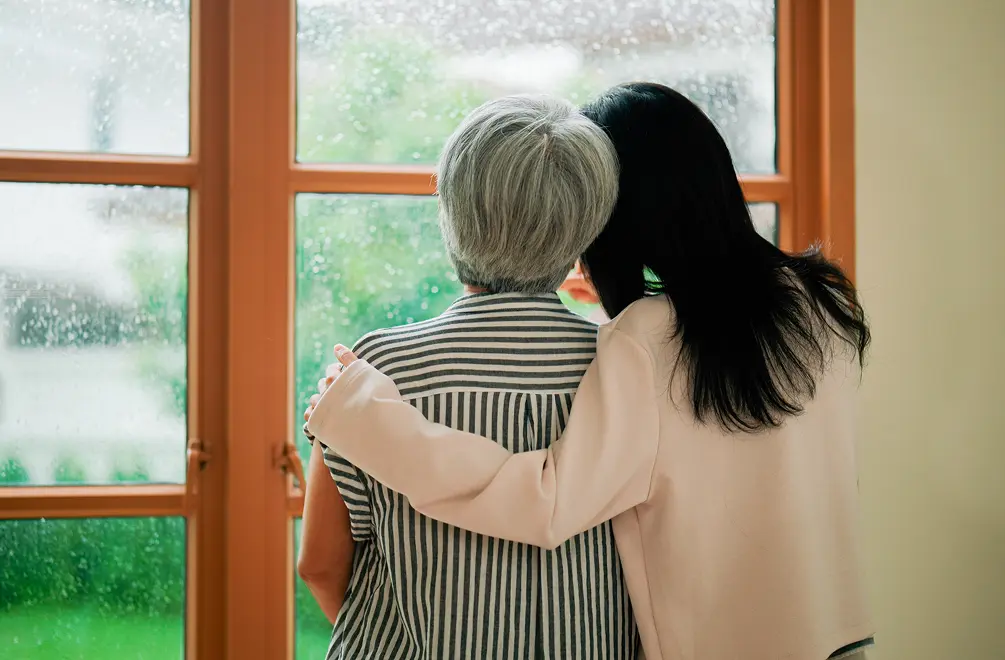 Two women, one elderly and one younger, embrace while looking out a rain-covered window.