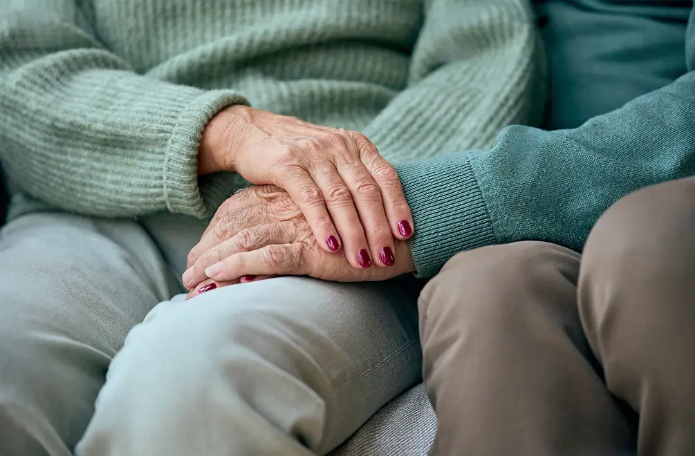 Close-up of two elderly people sitting side by side, one person gently holding the other's hand with painted fingernails.