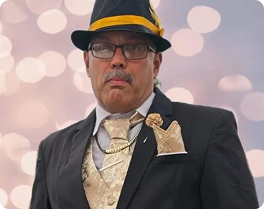 Man wearing a black suit with a gold patterned vest and tie, accessorized with a black hat featuring a gold band and flower, and glasses.