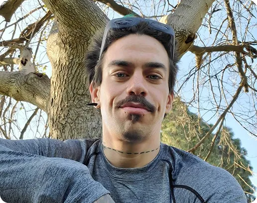 Man with a mustache and goatee wearing gray athletic shirt and sunglasses on his head, sitting outdoors near a tree.