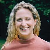 Smiling woman with curly blonde hair wearing a pink top, outdoors with green foliage background.