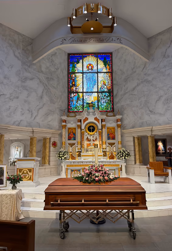 Interior of a marble church altar with a wooden coffin on a metal stand adorned with pink flowers in front. The altar features religious statues, floral arrangements, and a stained glass window depicting a radiant figure with a halo. Above the altar arches the phrase 'Gloria in Excelsis Deo.'