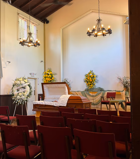 Interior of a chapel set up for a funeral service with a partially open wooden casket, floral arrangements of white and yellow flowers, rows of red chairs, and two chandeliers hanging from a high ceiling.