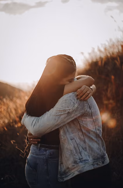 Two people embracing outdoors in a warm backlit setting, one wearing a blue denim jacket and the other with long dark hair, surrounded by tall grasses.