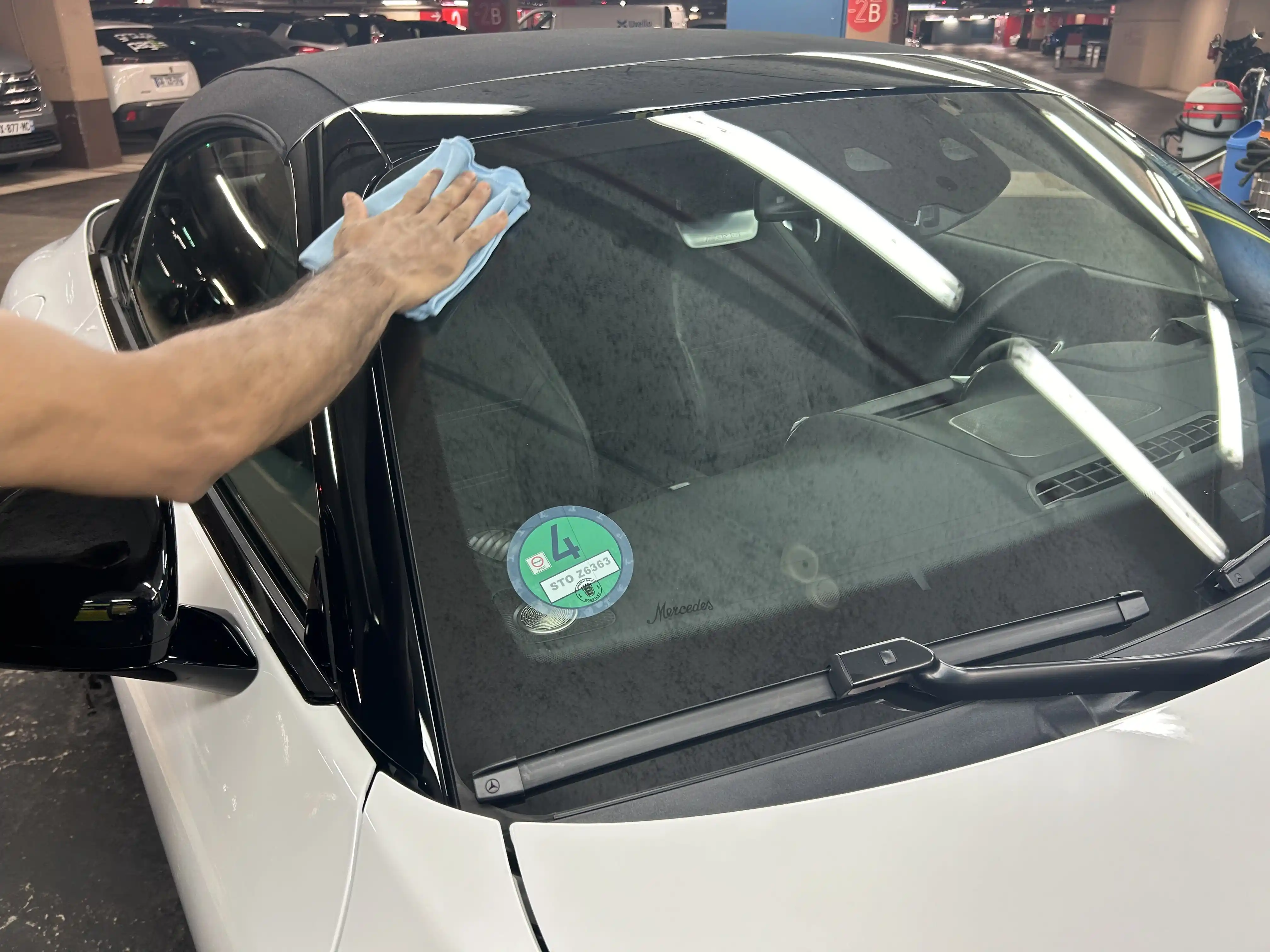 Une main essuie le pare-brise d'une voiture blanche dans un parking souterrain.