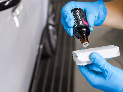 Personne portant des gants bleus versant de la céramique d'un petit flacon noir sur un morceau de tissu blanc à côté d'une voiture blanche.