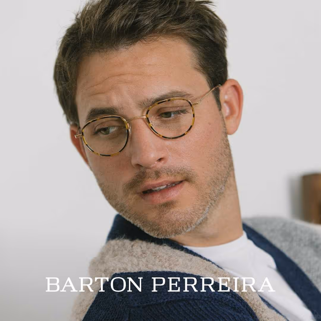 A man wearing barton perreira glasses and a sweater looks at the camera