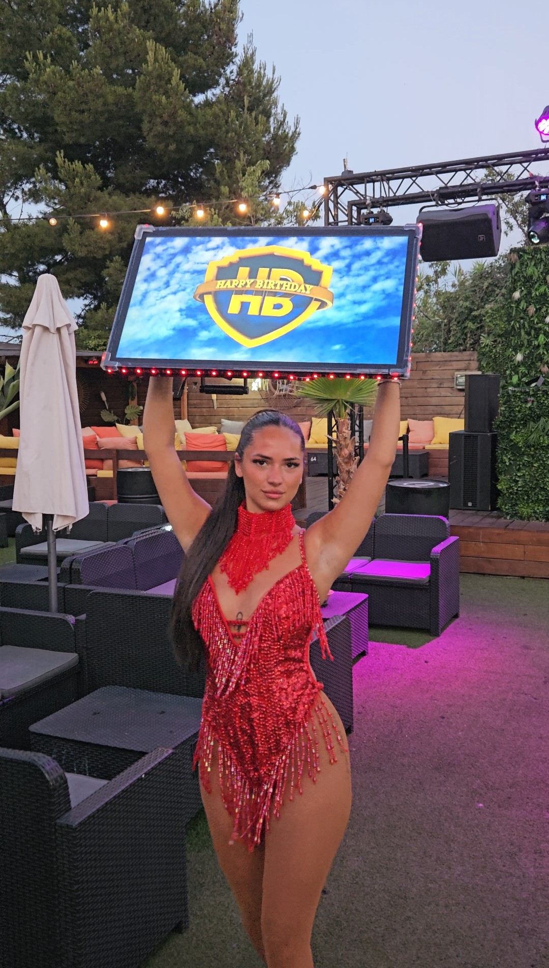 Jeune femme en costume rouge pailleté tenant un panneau lumineux avec l'inscription Happy Birthday dans un cadre extérieur lounge.