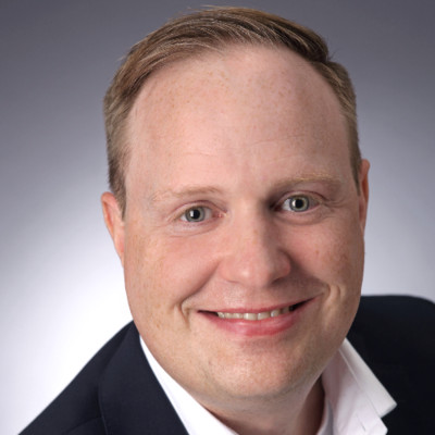 Smiling man with light hair wearing a dark jacket and white shirt against a gray background.