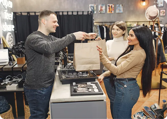 Man handing a shopping bag to a smiling woman in a clothing and accessories store with another woman nearby.