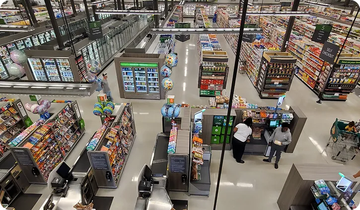 View of a supermarket checkout area with aisles stocked with groceries and two people at the counters.