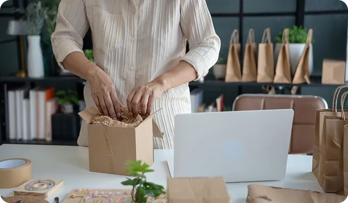 Person packing items into a cardboard box on a desk with a laptop and brown paper bags nearby.