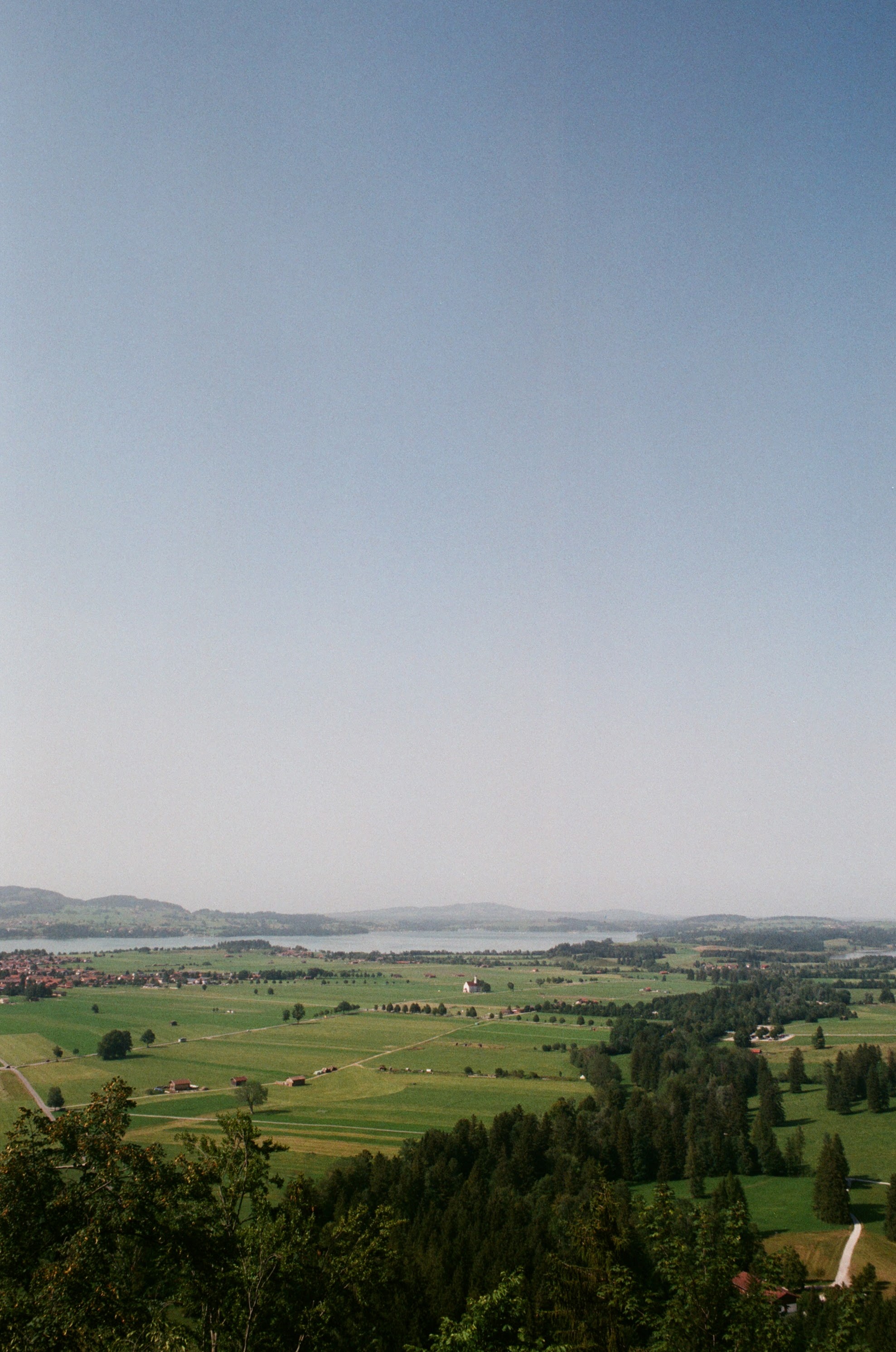Pastoral landscape of Bavarian countryside with rolling green fields, scattered farms, and distant lake beneath soft gray skies