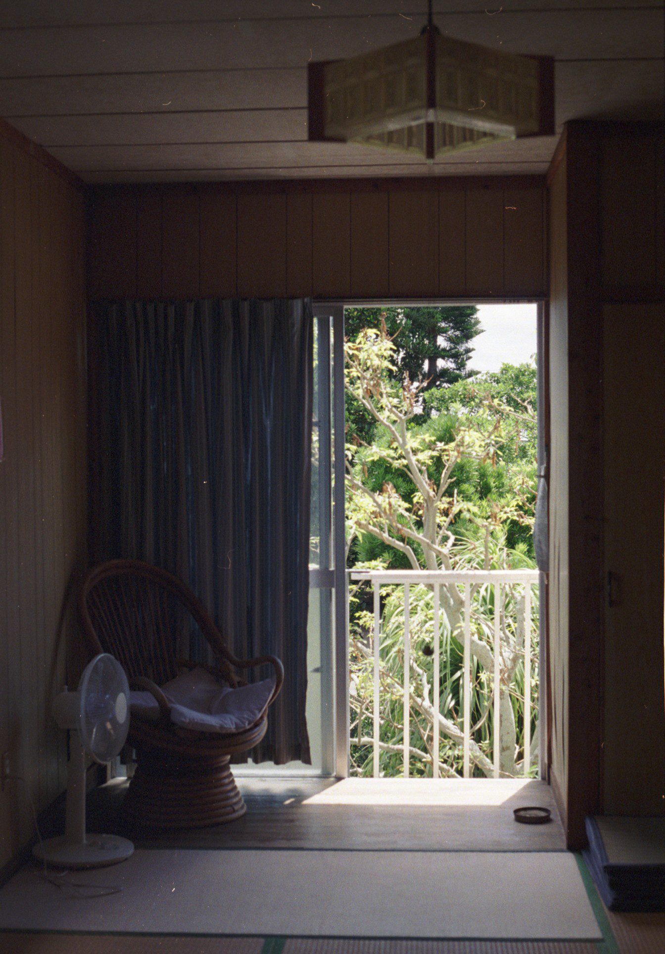 English: “Kuroshima, Okinawa, Japan: a dim wooden room with tatami flooring, a wicker chair, and a small fan, looking out through an open sliding door to a sunlit balcony and dense greenery.”

日本語: 「沖縄県黒島：薄暗い木造の部屋に畳と籐椅子、扇風機があり、開いた引き戸の先に日差しのバルコニーと濃い緑が見える。」