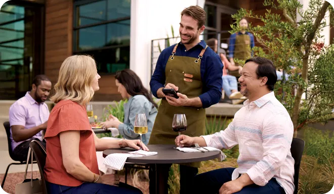 Man waiter in apron taking order from a smiling couple seated at an outdoor restaurant table.