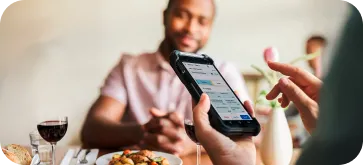 Person holding smartphone showing a food delivery app at a dining table with a man in the background