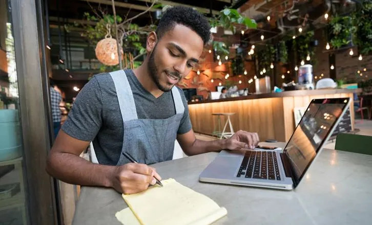 Man in apron writing on notepad while using laptop at a café table with warm lighting.