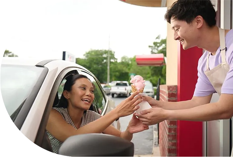 Woman in car receiving ice cream cone from smiling server at drive-thru window.