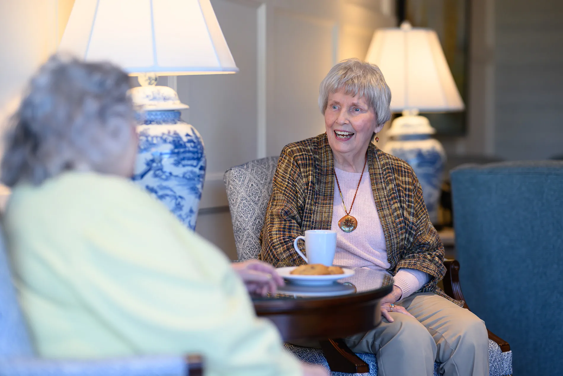 Two elderly women sitting and chatting in a cozy room, one smiling and holding a cup at a small table with cookies.