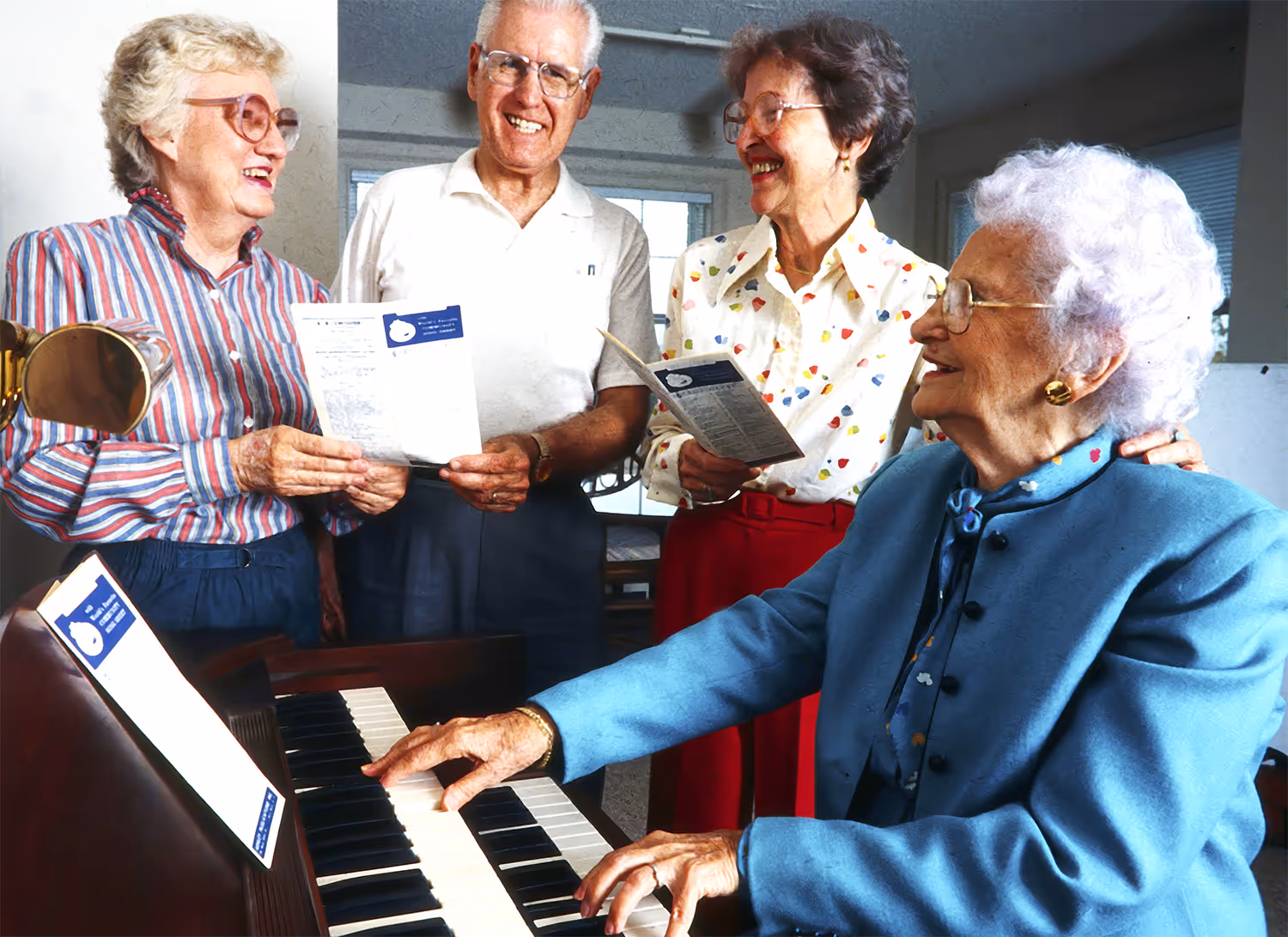 Four elderly people smile and sing together while one plays the piano.