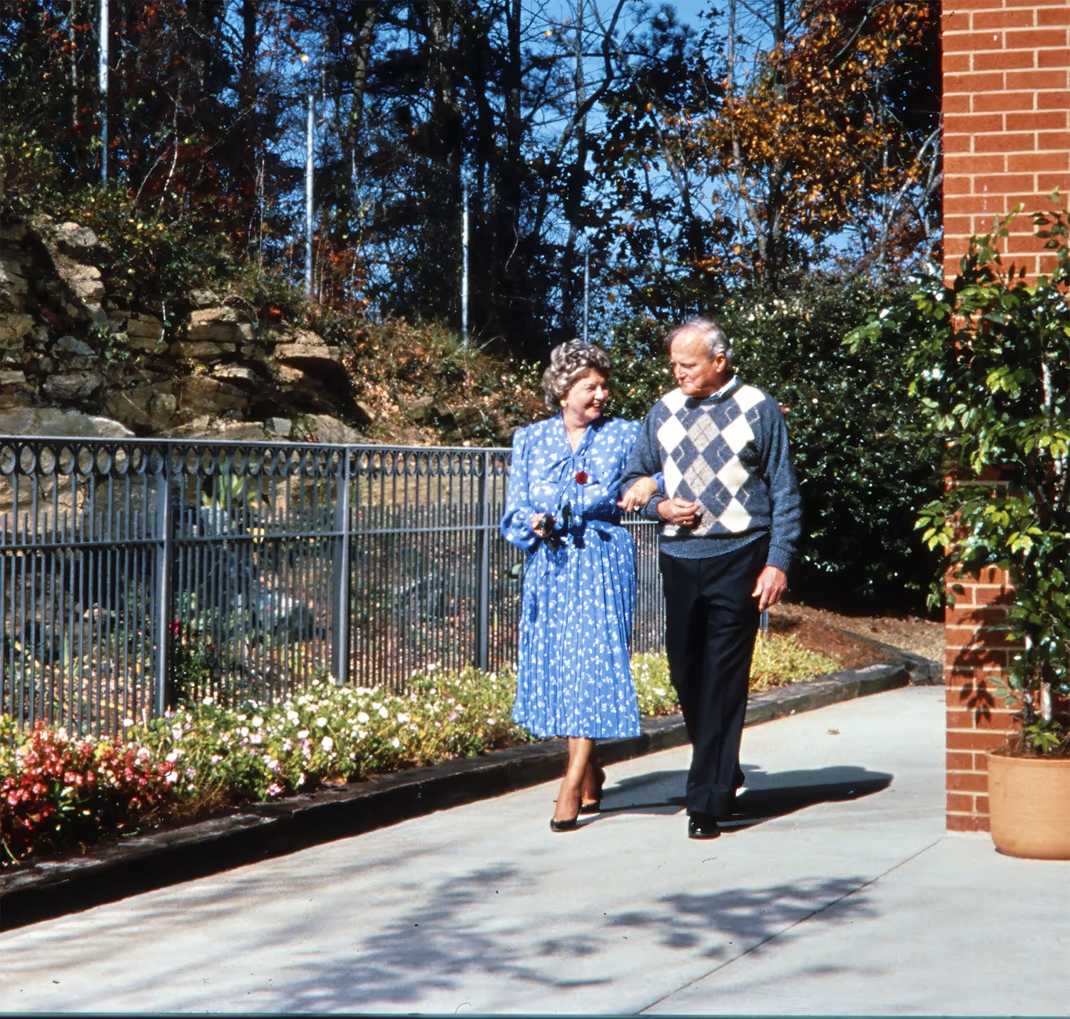 An elderly couple walks together on a garden path, holding hands and smiling.
