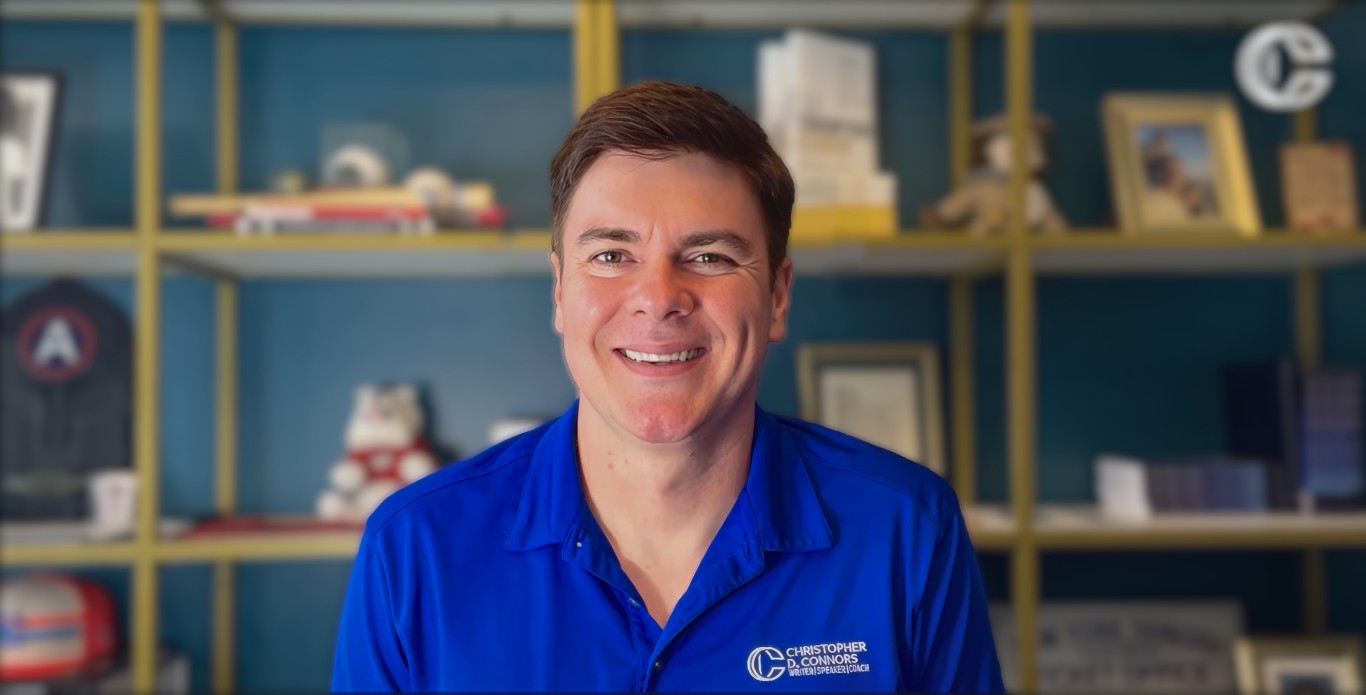 Smiling man wearing a blue shirt with Christopher D. Connors logo, in front of a bookshelf with various items.