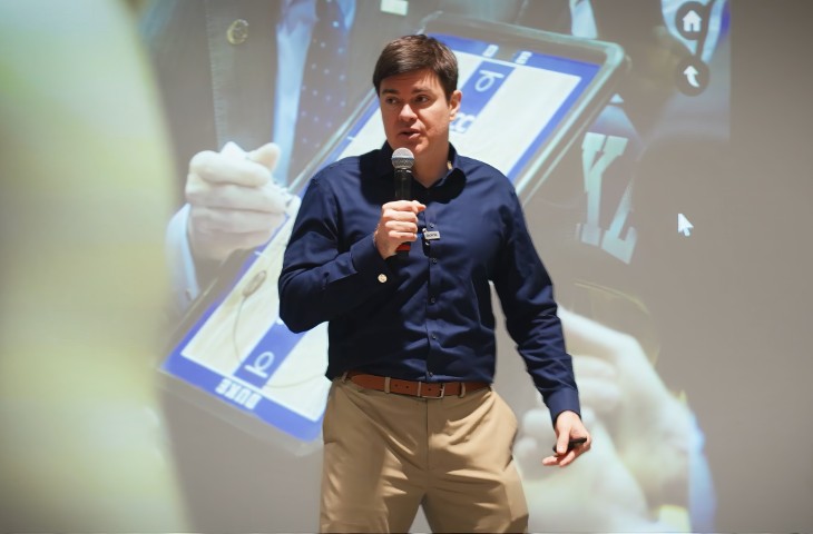 Man in navy blue shirt and khaki pants speaking into a microphone during a presentation with a large screen behind him.
