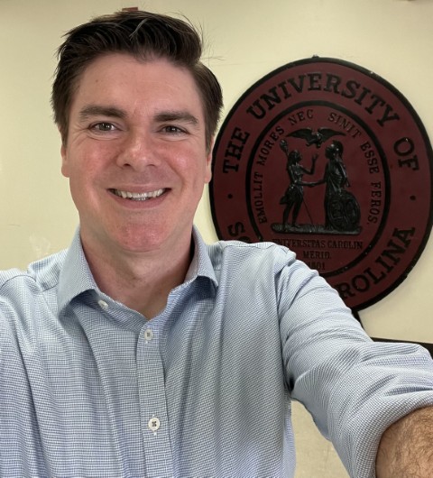 Christopher D. Connors selfie in front of a University of South Carolina seal on a beige wall.
