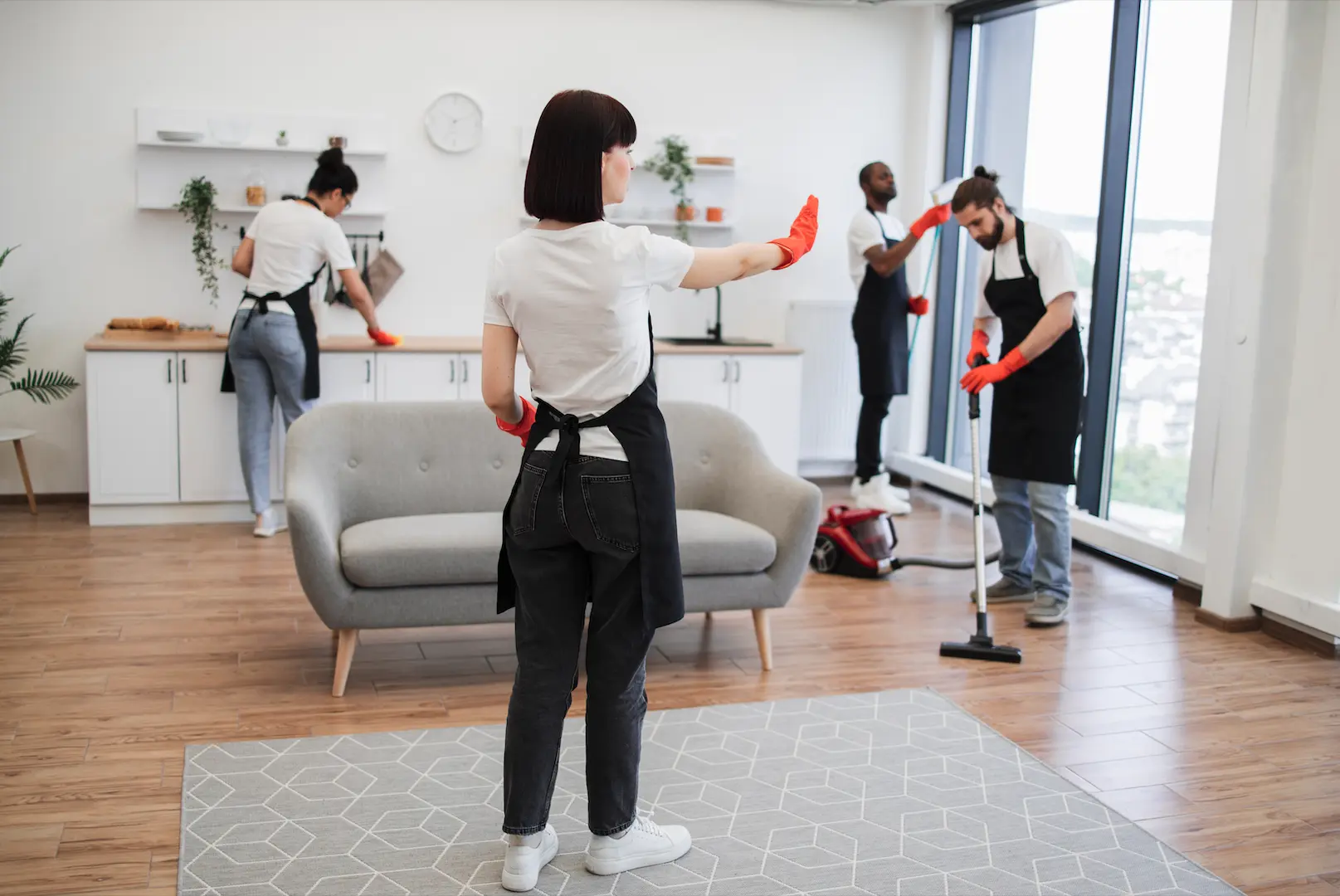 Cleaning team wearing aprons and orange gloves tidying a modern living space with sofa and large windows.