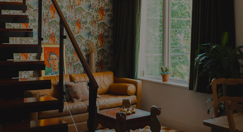 A decorative room with a comfortable leather couch and stylish checkered run. The wall is decorated with ornate floral patterned wallpaper and tasteful floor-to-ceiling emerald green curtains.