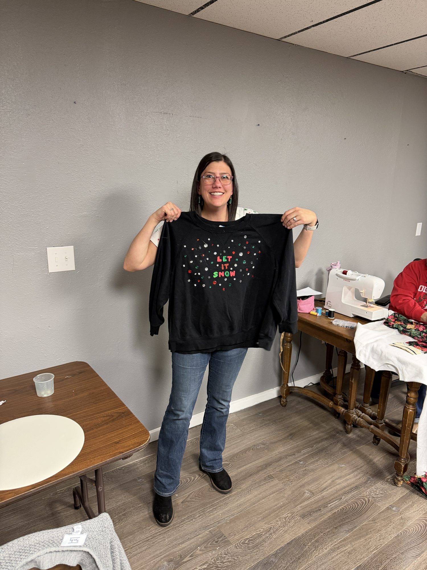 A smiling woman holds up a black sweater with sparkly details.