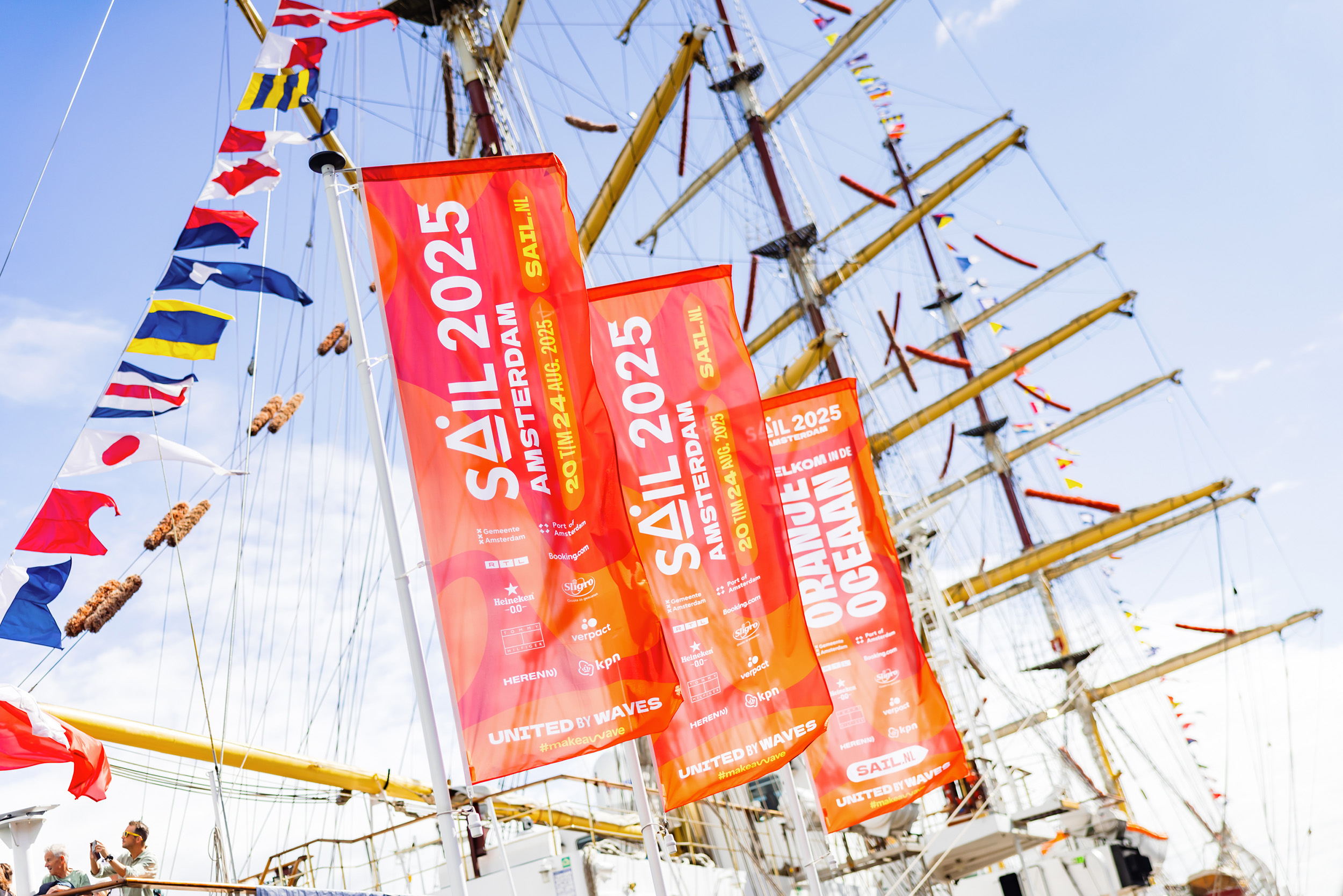 Vlaggen in de huisstijl van SAIL 2025, met op de achtergrond de masten van een tall ship. 