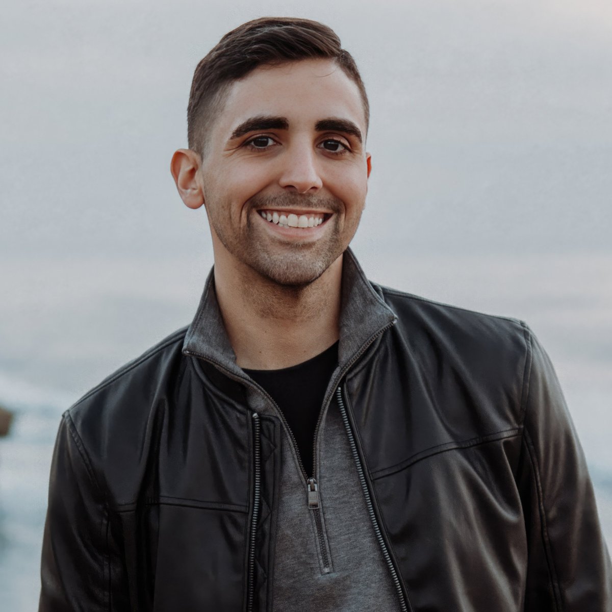 Smiling young man with short dark hair wearing a black leather jacket over a gray zip-up shirt, standing outdoors with a blurred light background.