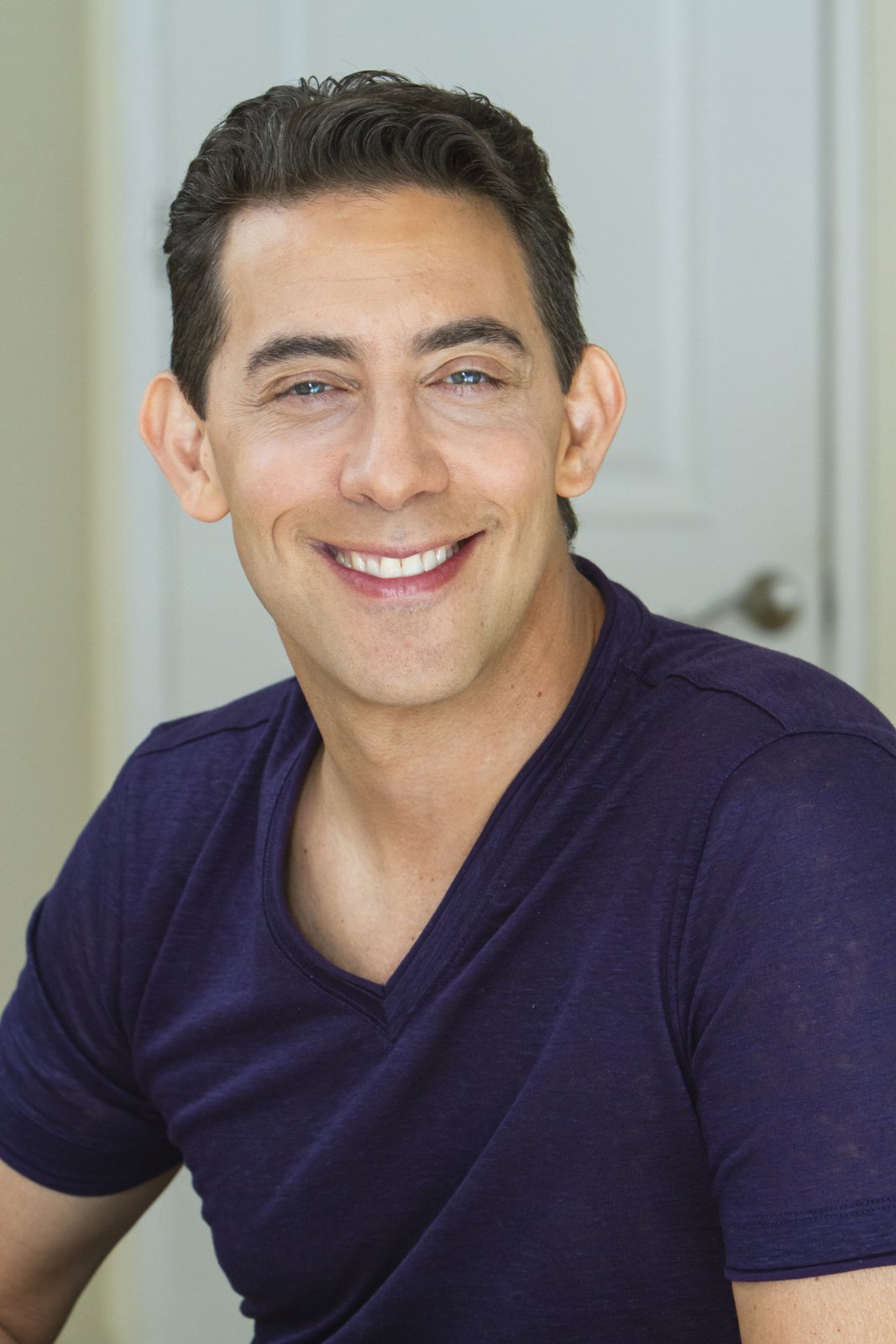 Smiling man with short dark hair wearing a purple V-neck shirt, seated indoors with a blurred background.