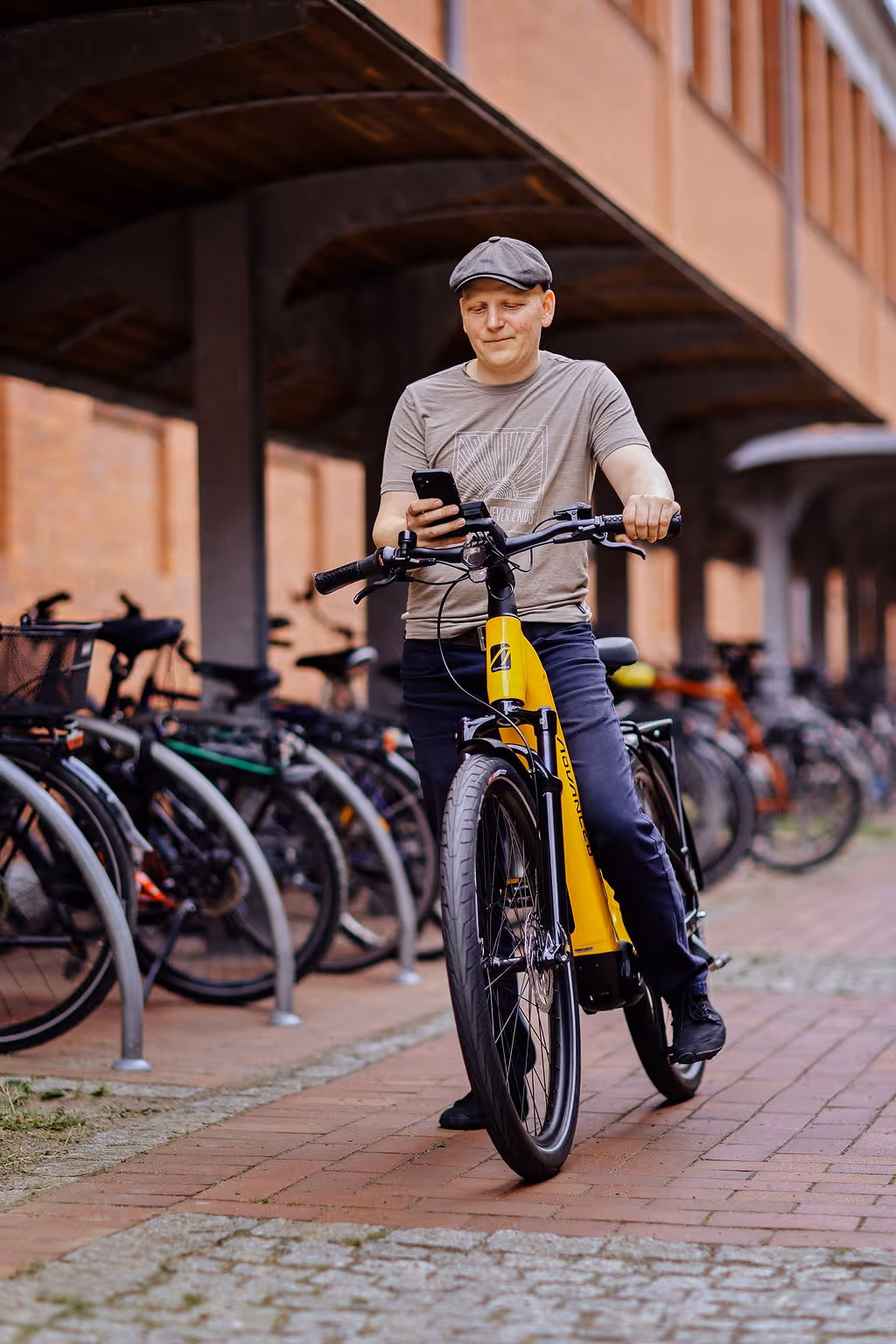Mann mit grauem Shirt und Schirmmütze steht mit gelbem Elektrofahrrad und schaut auf sein Smartphone.