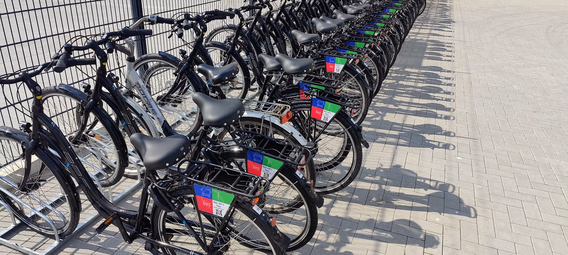 Reihe von schwarzen Fahrrädern zum Ausleihen mit Werbeschildern auf gepflastertem Boden.