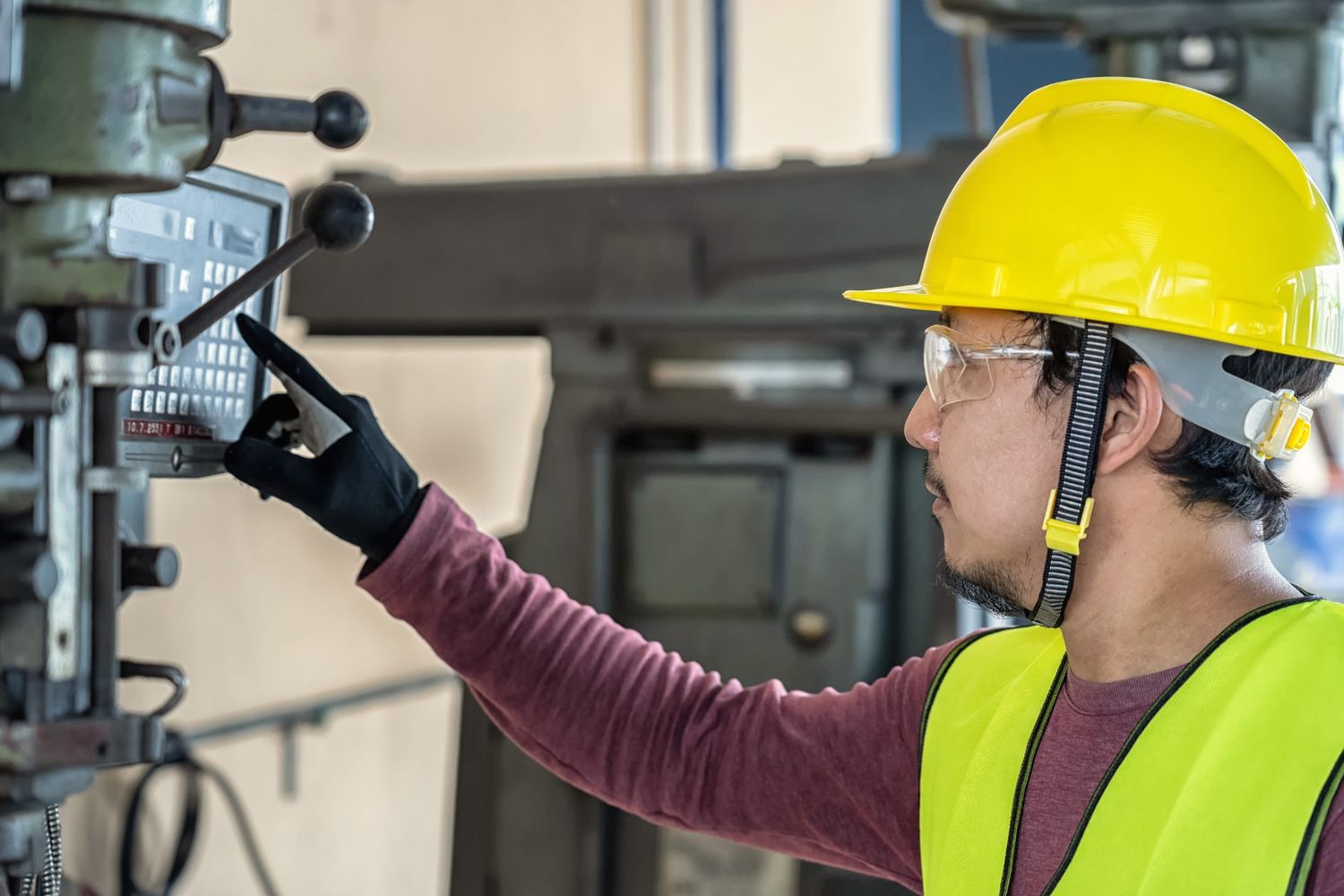 A Worker wearing a yellow hard hat and a high-visibility vest operates machinery in an industrial setting, focusing on controls with their gloved hand.

