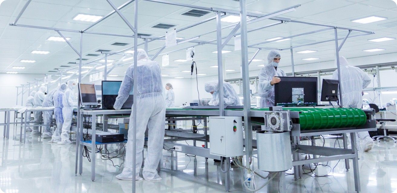 A bright, clean room with several people in lab coats and protective gear working at various stations, possibly in a manufacturing or research facility. The environment suggests a sterile or controlled setting.