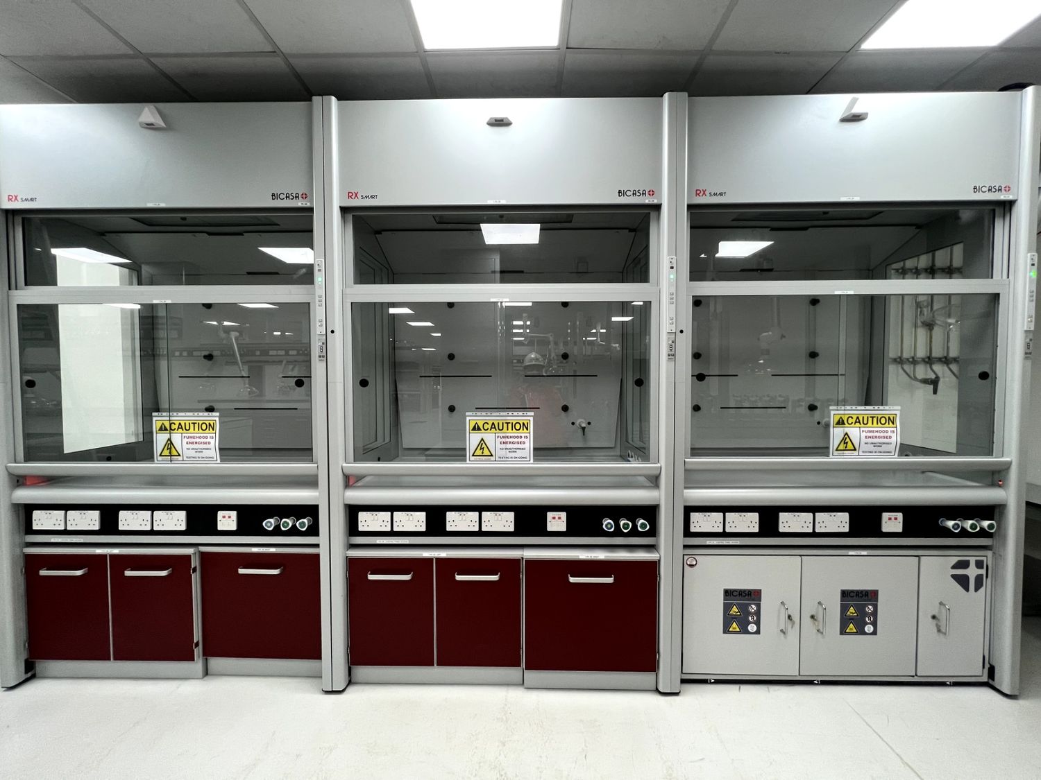A line of three empty laboratory fume hoods with safety warning signs and red cabinets.