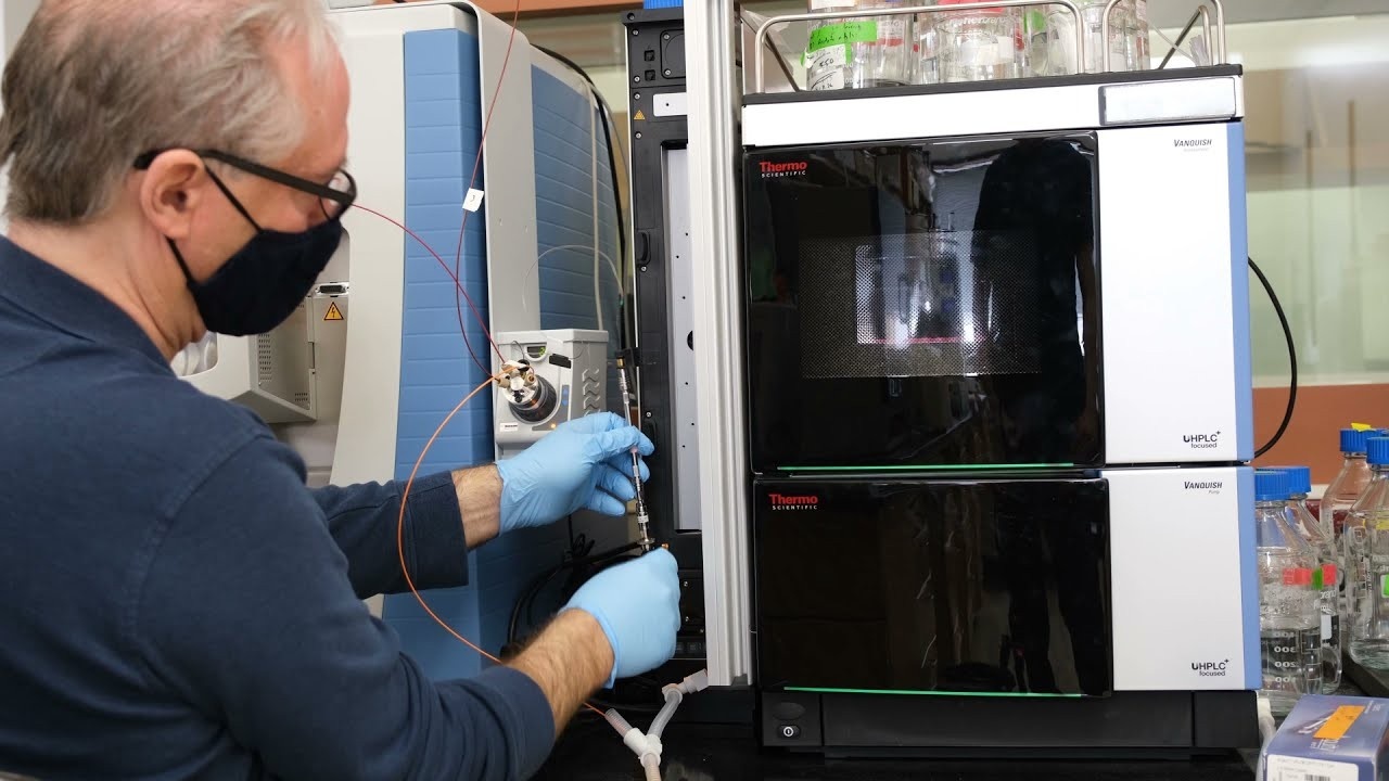 A masked scientist in blue gloves working on a large analytical instrument in a laboratory.