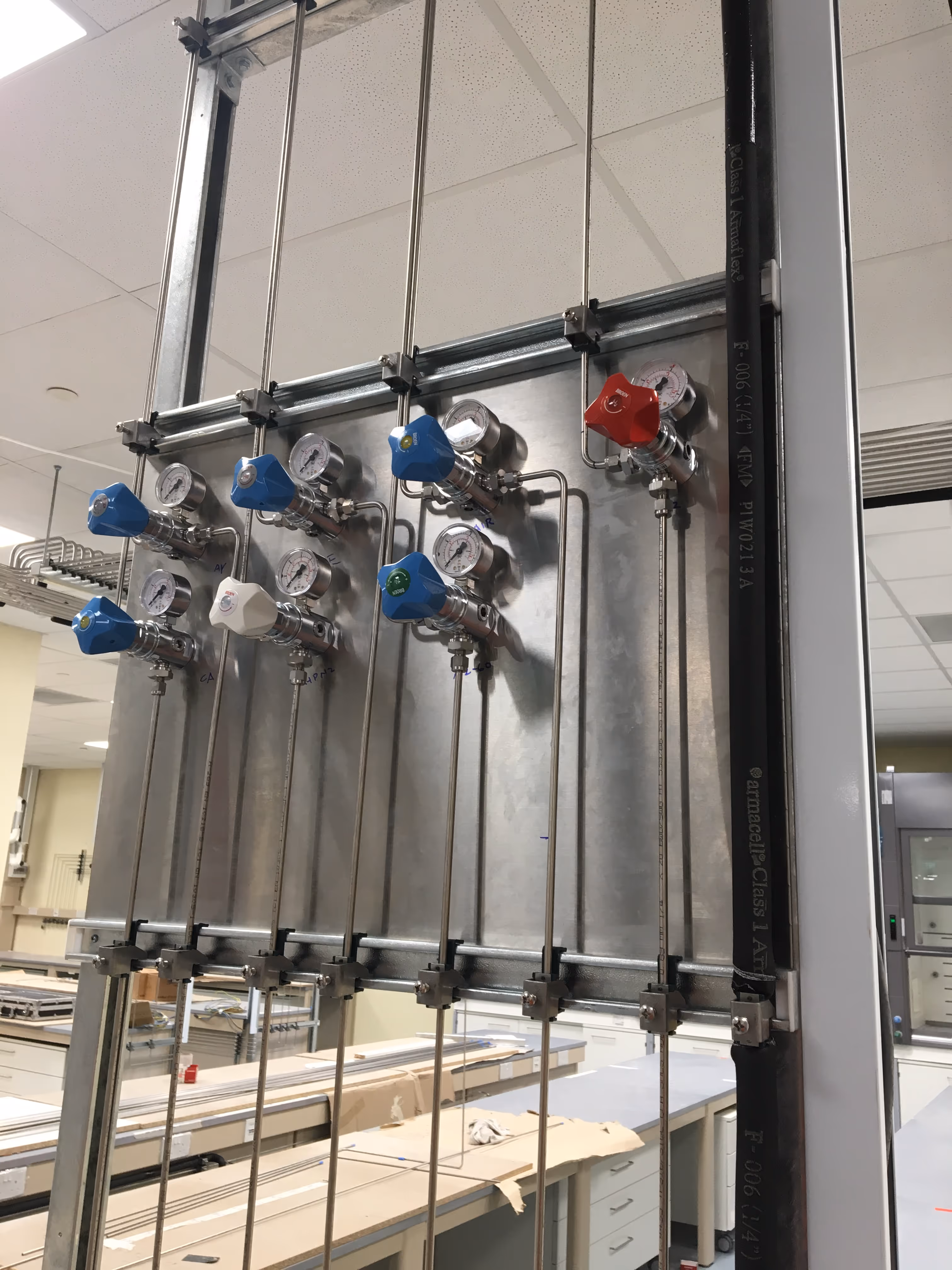 Close-up of a wall-mounted laboratory gas control panel featuring multiple stainless steel lines, pressure gauges, and color-coded manual regulators  in a lab environment.