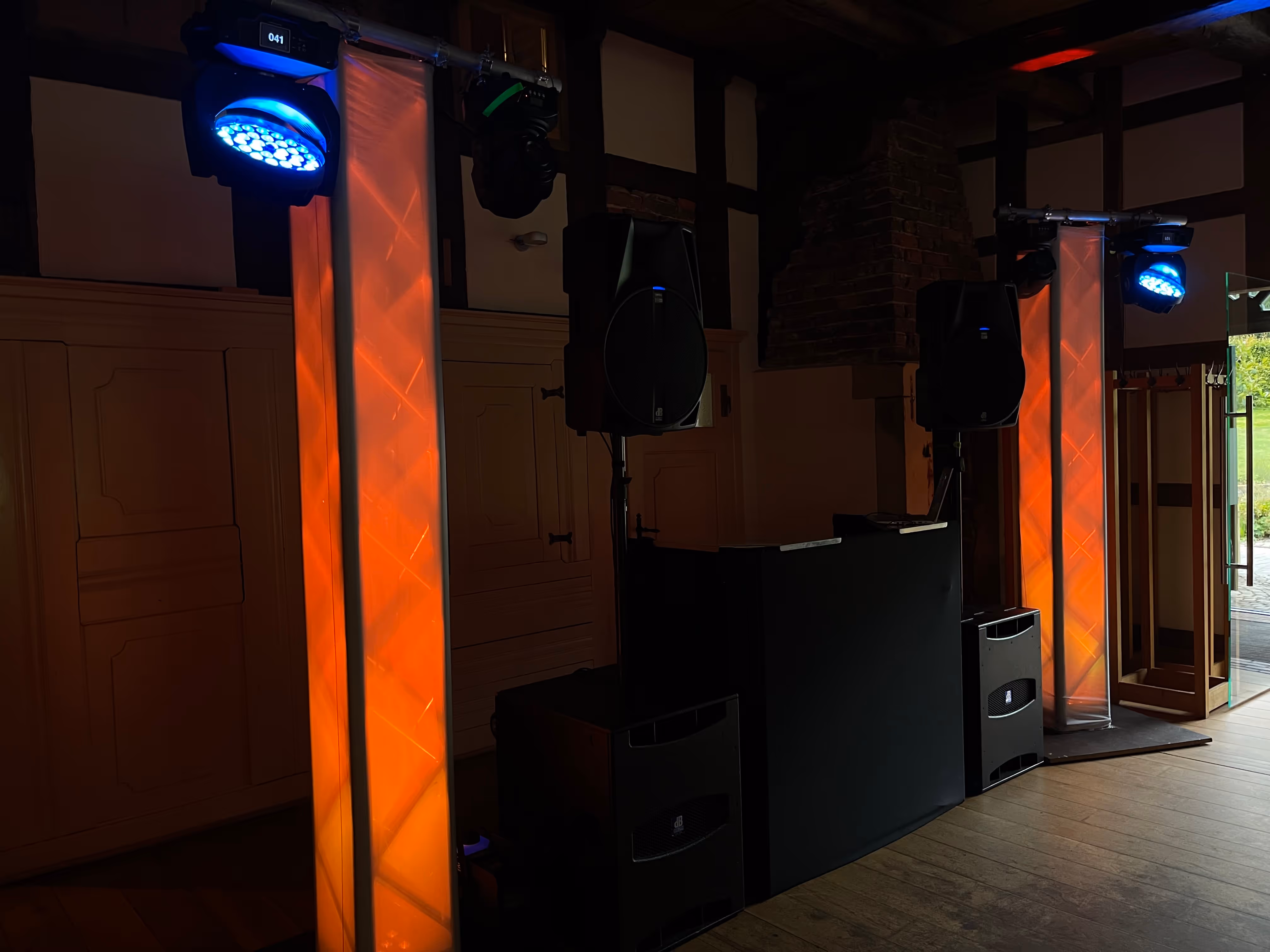 Indoor-DJ-Setup mit Lautsprechern, schwarzem DJ-Pult und zwei vertikalen orange leuchtenden Lichtsäulen sowie blauen Scheinwerfern.
