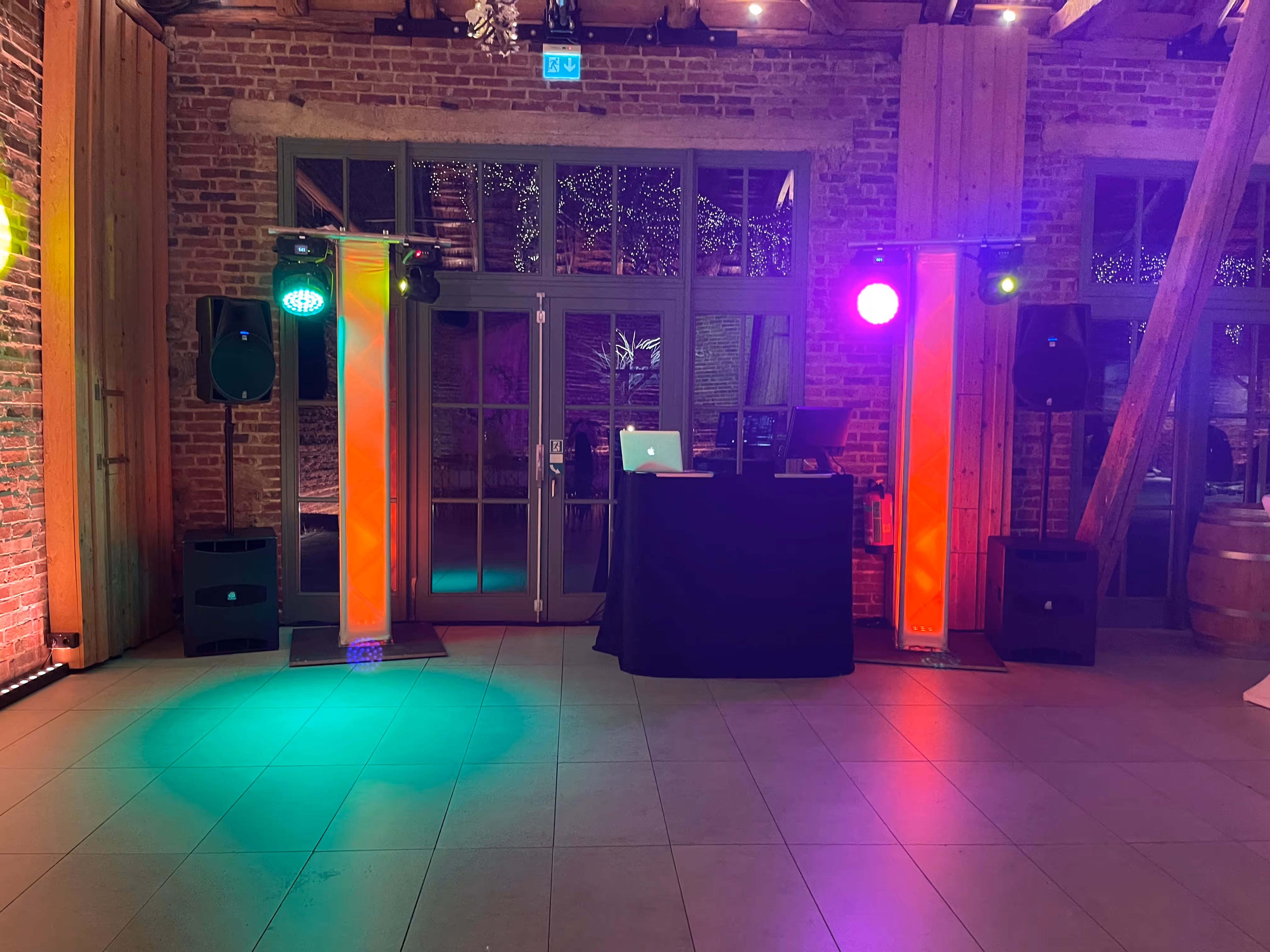 Indoor-Event-Setup mit DJ-Pult inklusive Laptop, Lautsprechern und bunten Scheinwerfern, die grünes, lila und rotes Licht auf den Fliesenboden werfen.