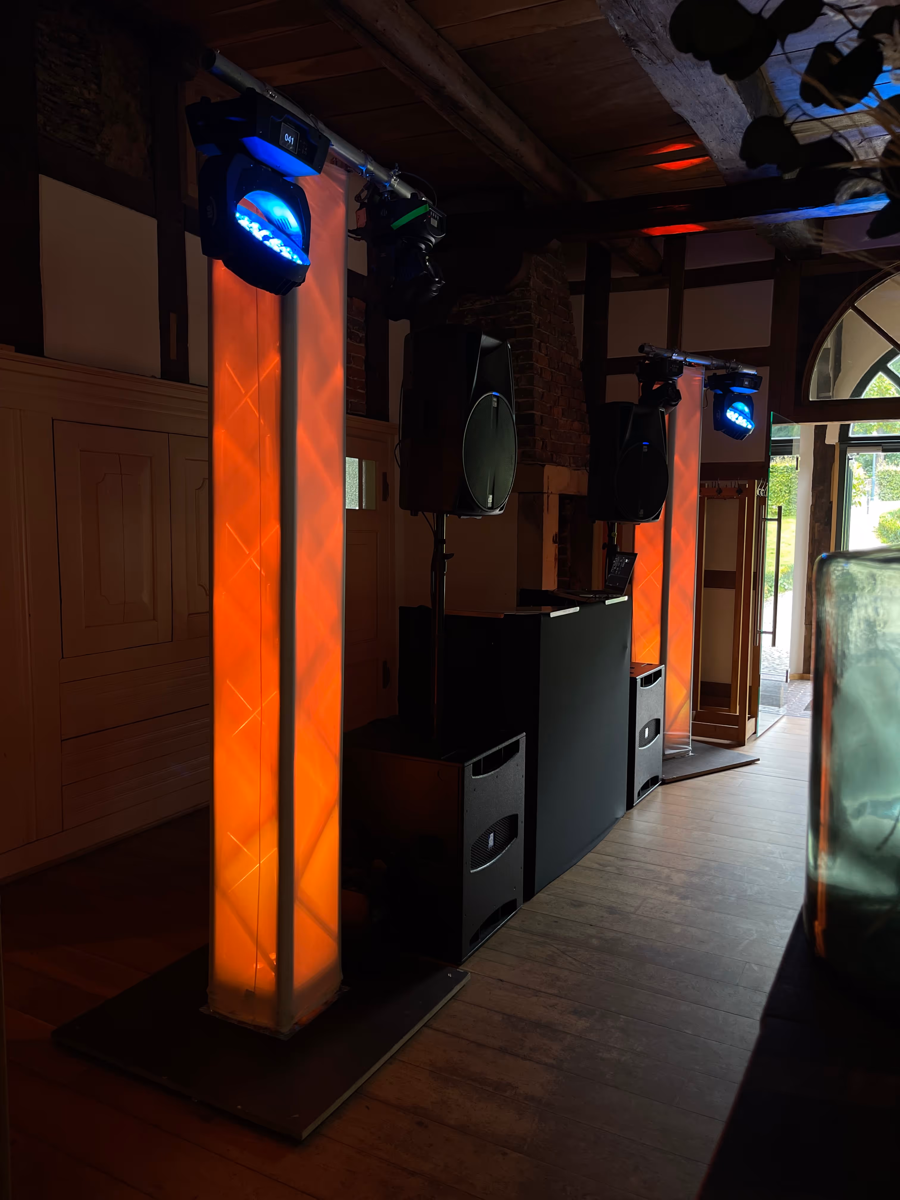Indoor-DJ-Setup mit schwarzen Lautsprechern und orange beleuchteten Lichtsäulen neben einem Holzboden und einer offenen Tür.