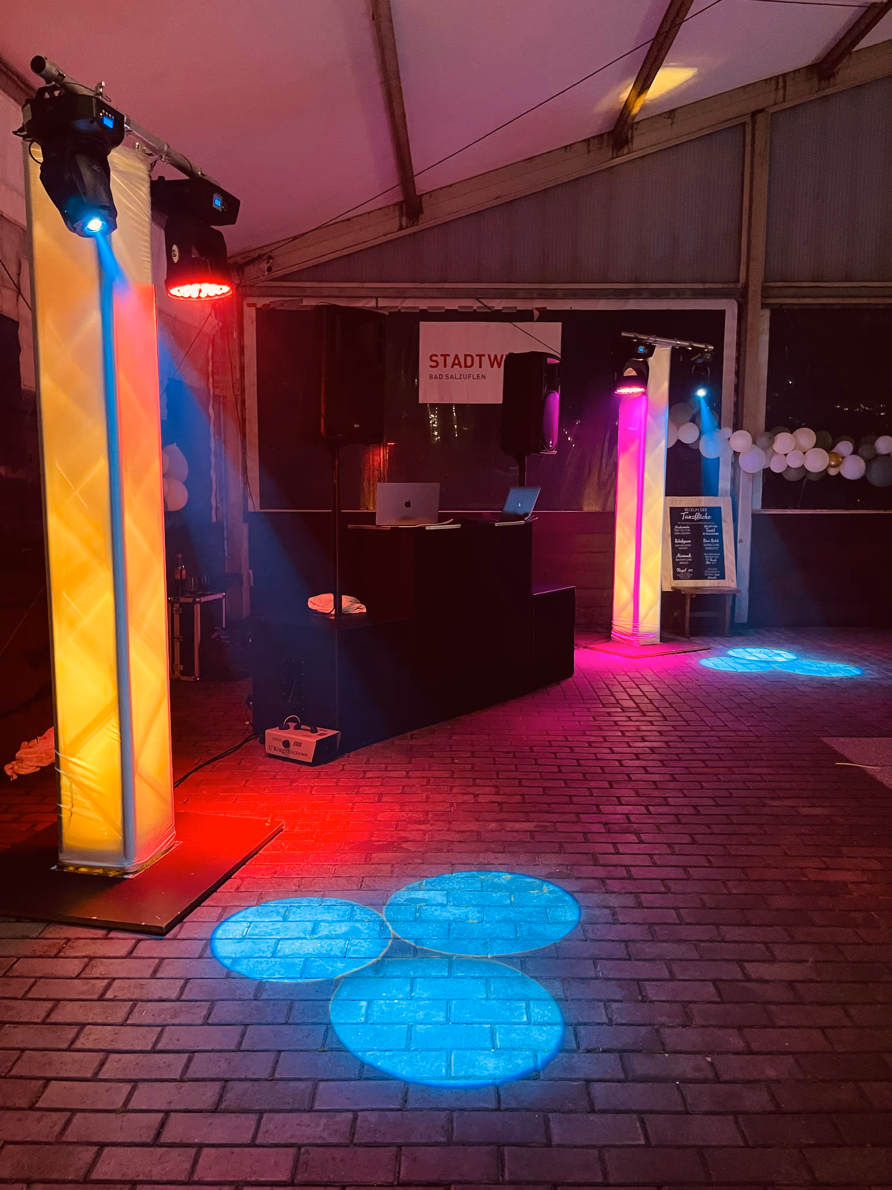 Indoor-Eventlocation mit beleuchtetem DJ-Pult, bunten Scheinwerfern und blauen Spotlights auf Backsteinboden.