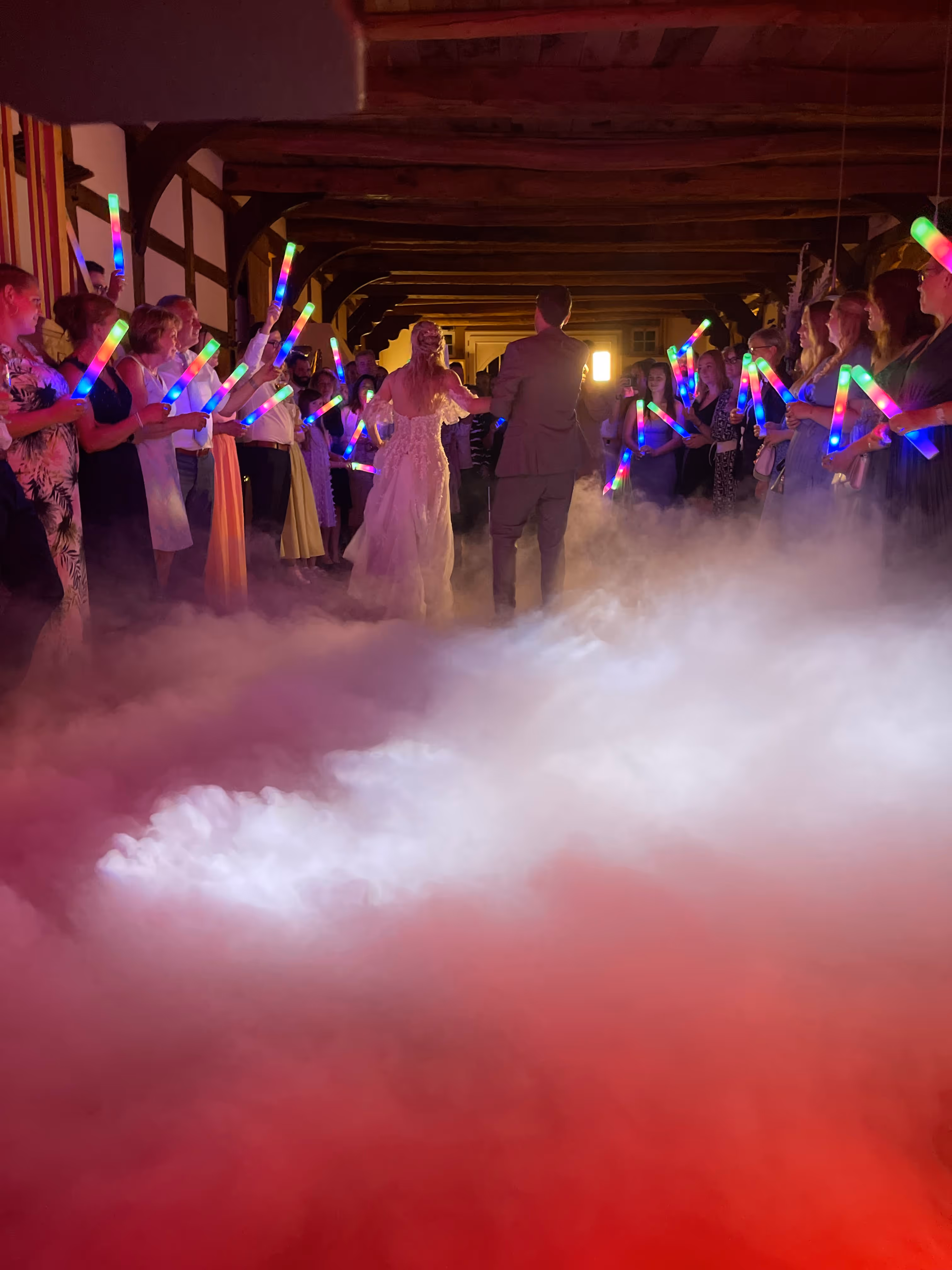 Brautpaar beim Tanzen in einer dekorierten Location mit Bodennebel und Gästen, die bunte Leuchtstäbe halten.