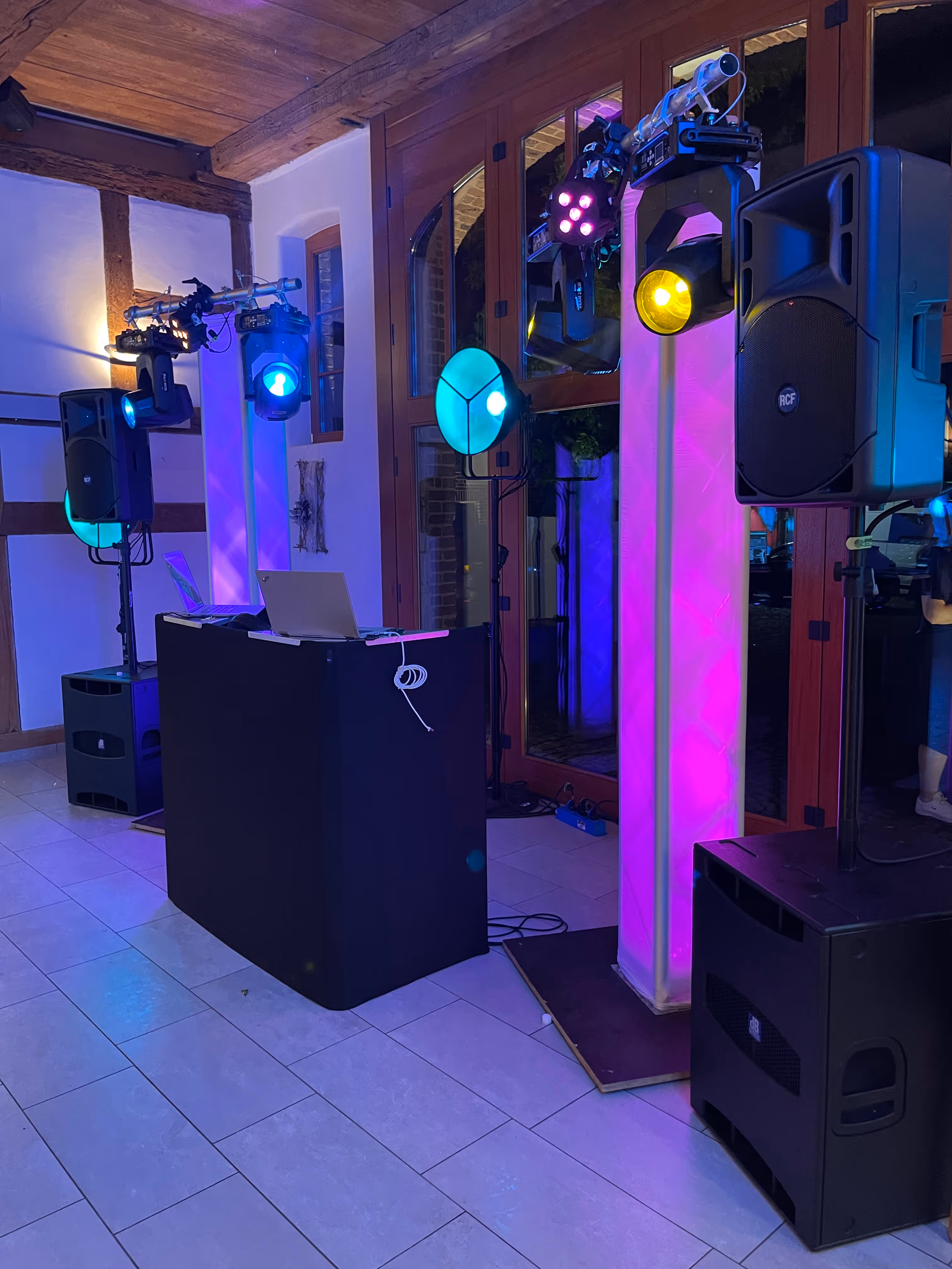 Indoor-DJ-Setup mit Laptop, schwarzen Lautsprechersystemen und bunten Scheinwerfern, die an und um beleuchtete weiße Säulen montiert sind.