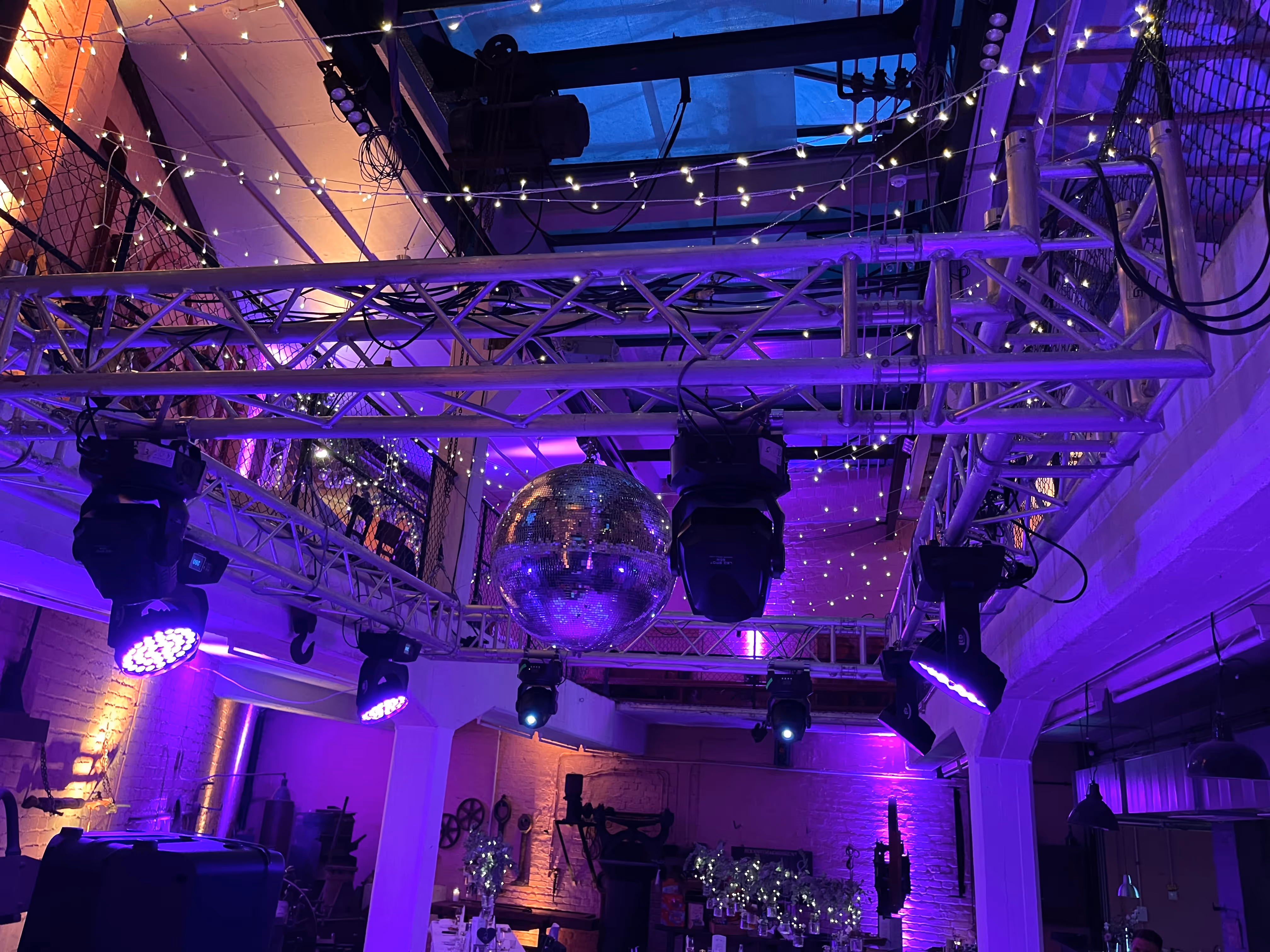 Indoor-Eventlocation mit lila Beleuchtung, einer an Traversen hängenden Discokugel und Lichterketten an der Decke.