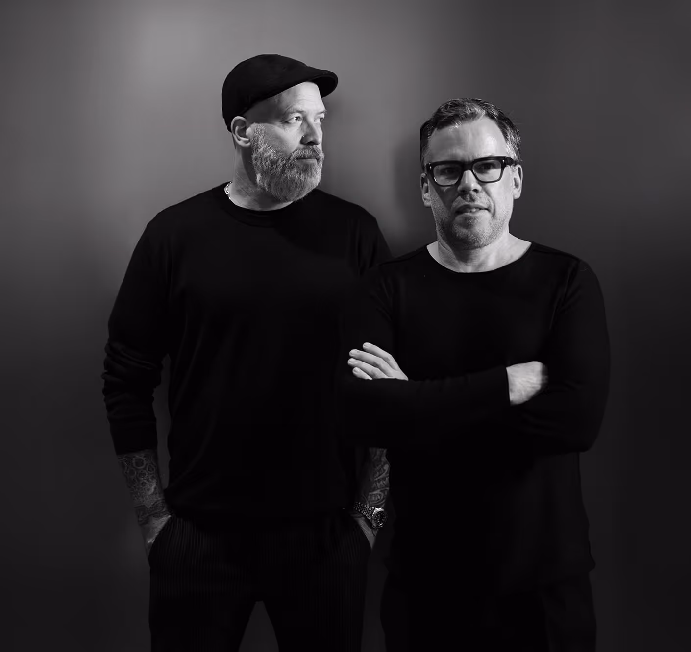 Black and white portrait of two men standing side by side against a plain background, one with a beard and cap looking left, the other wearing glasses with arms crossed.